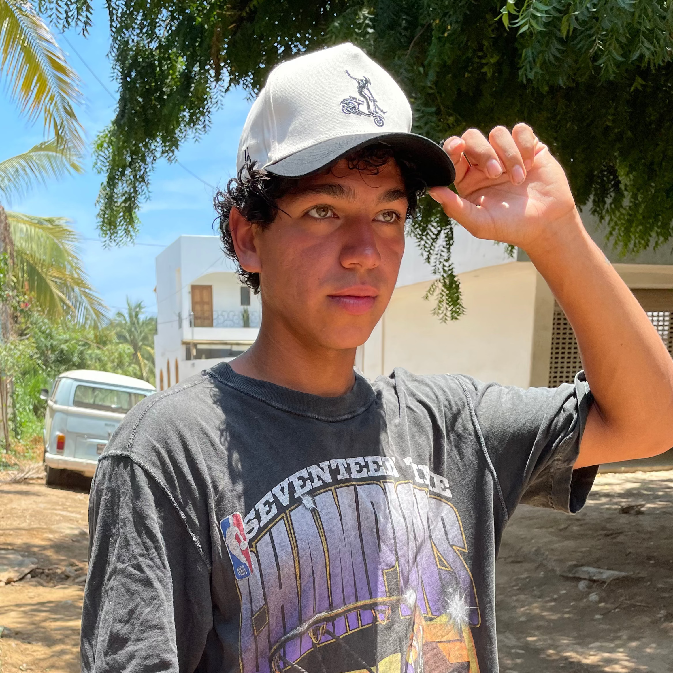 Person wearing a graphic t-shirt and cap standing next to a scooter outdoors.